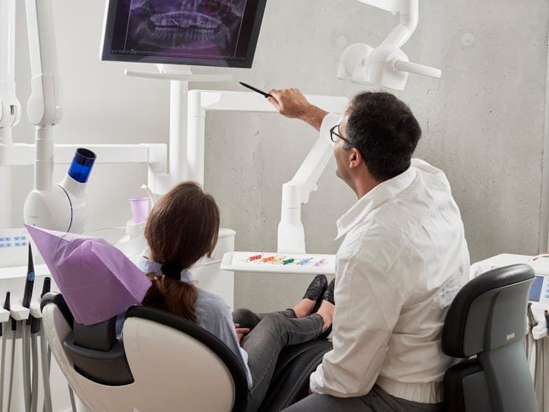 Cirurgião-dentista apontando a radiografia panorâmica na tela para a paciente na cadeira