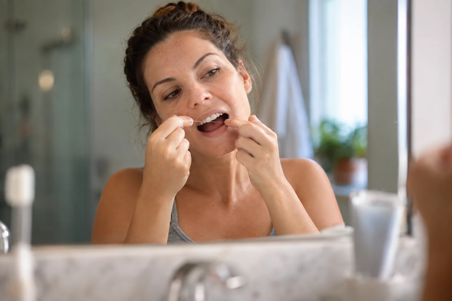 Mulher passando fio dental em frente ao espelho do banheiro pela manhã.